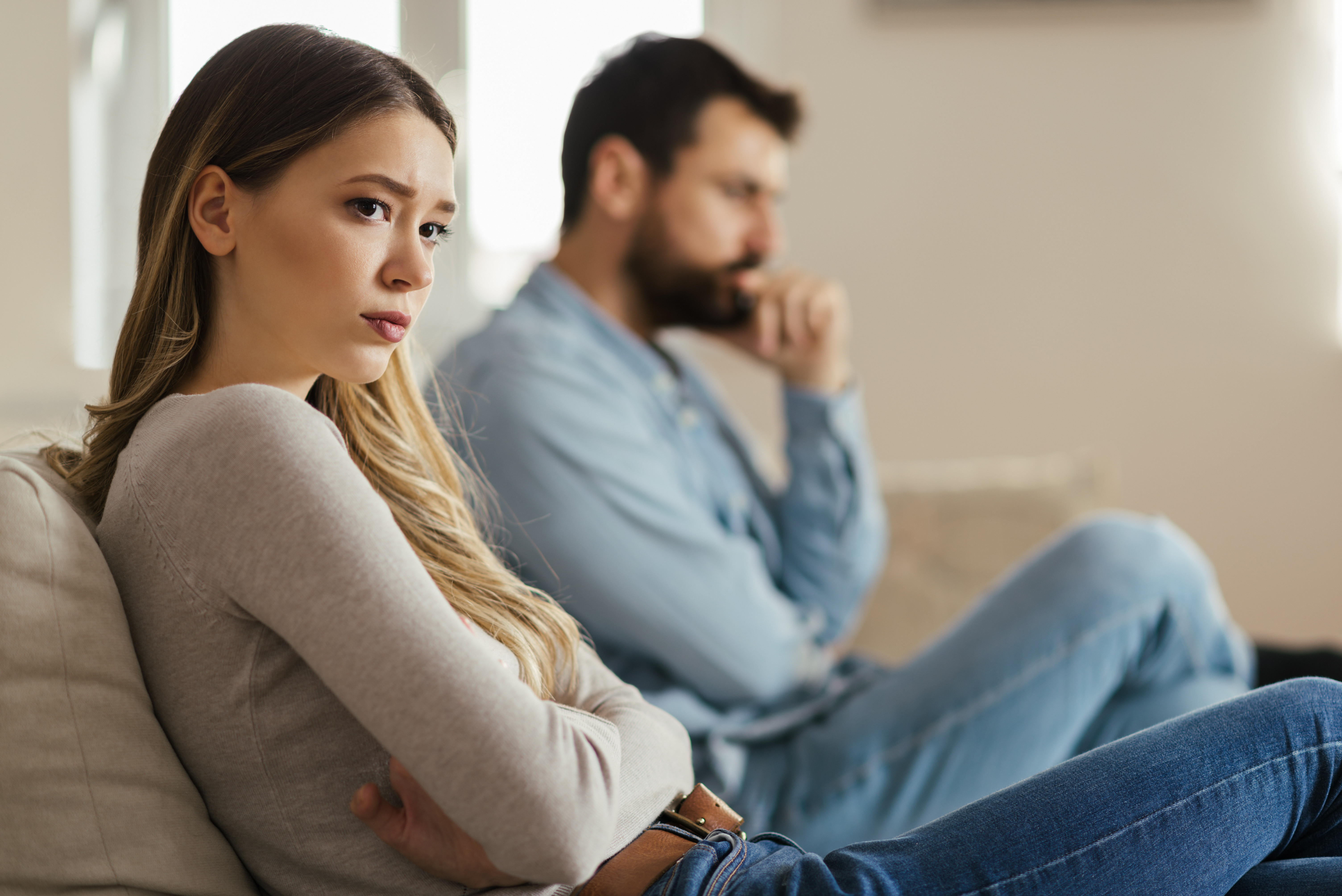 Worried young woman sitting on sofa at home and ignoring her husband who is sitting next to her