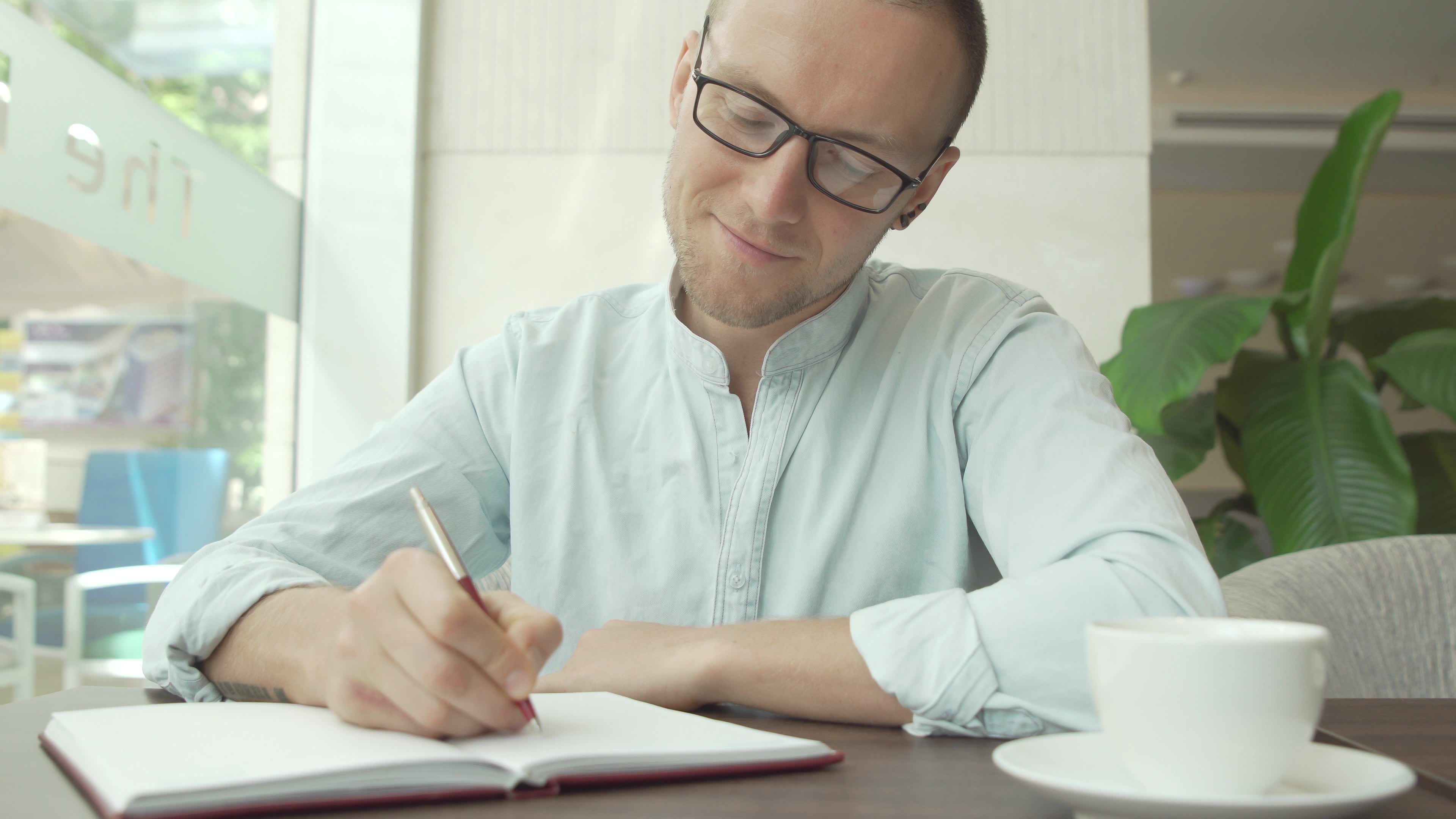Young man with glasses making notes in a journal while he drinks coffee