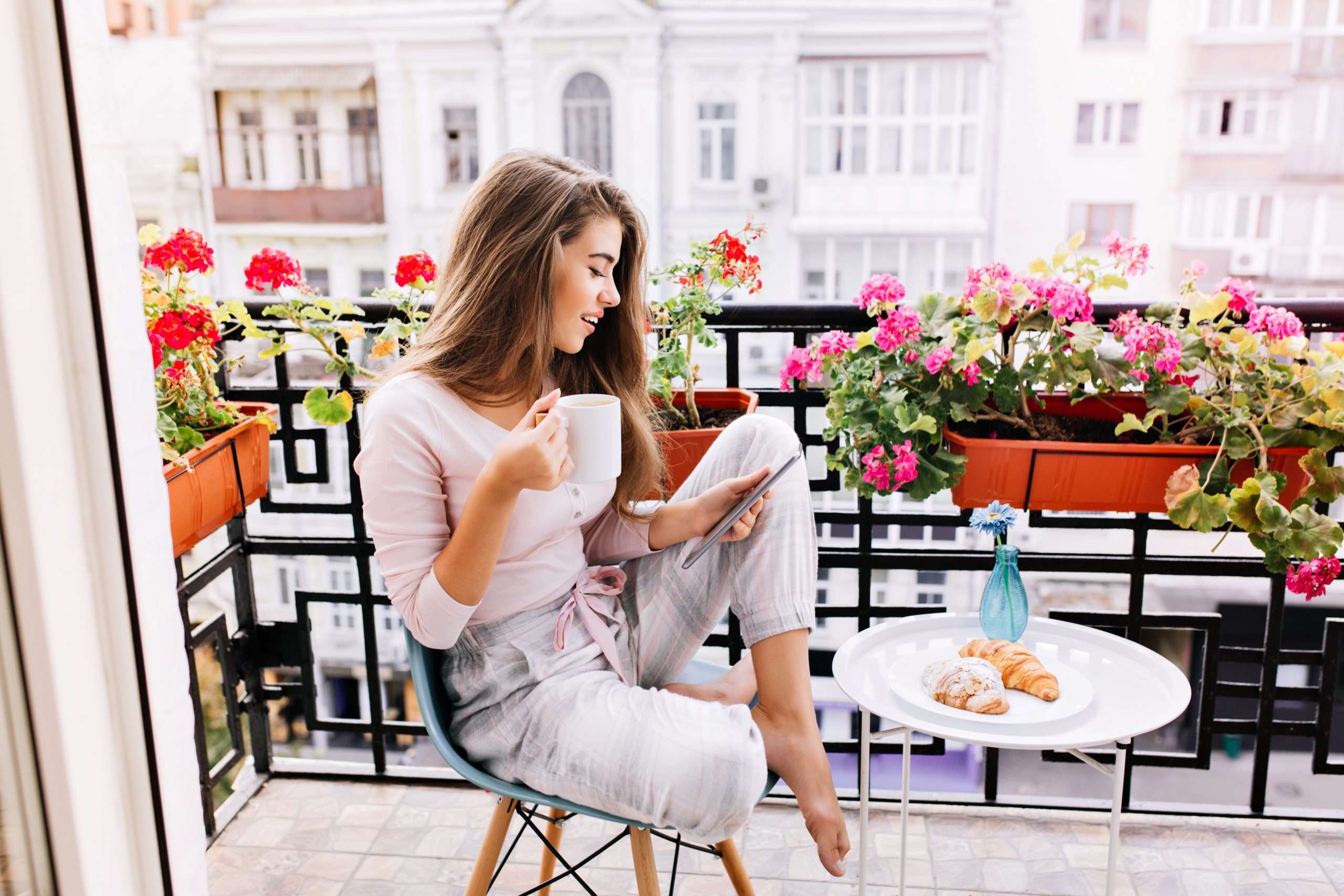 Attractive girl with long hair in pajama having breakfast on balcony in the morning in city. She holds a cup, reading on tablet.