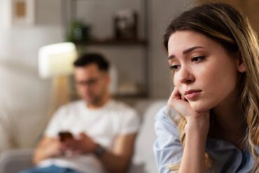 Upset woman sits on one end of couch while her boyfriend sits on the other end, looking at his phone