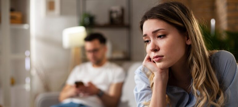 Upset woman sits on one end of couch while her boyfriend sits on the other end, looking at his phone