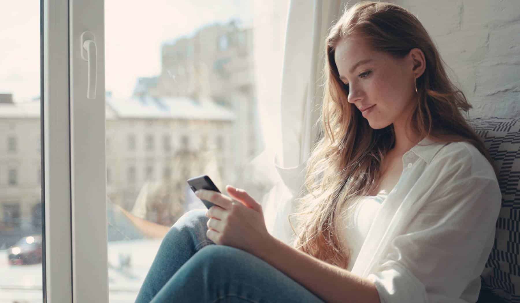 Young woman sitting on a windowsill at home and texting